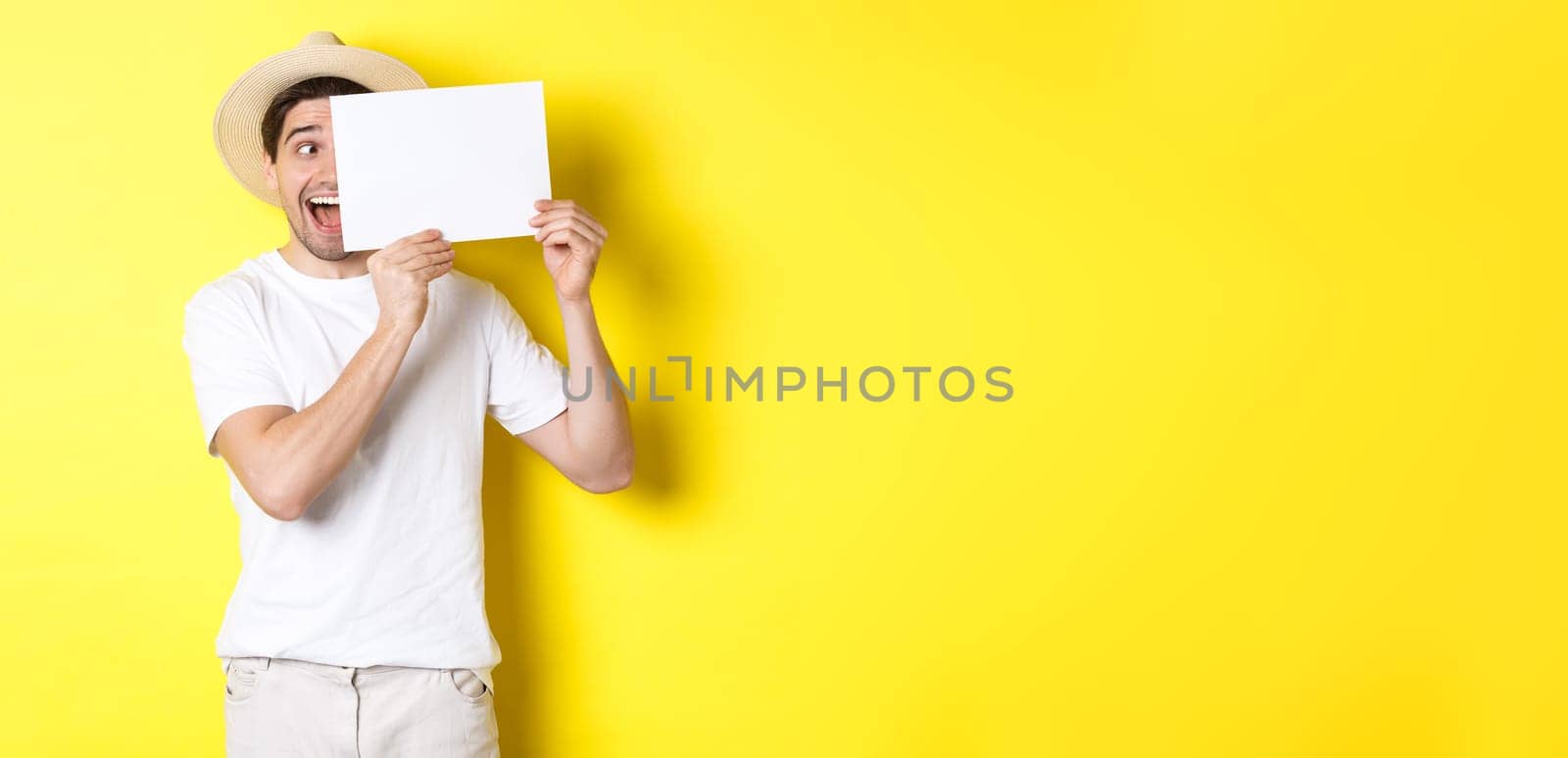 Excited man on vacation showing blank piece of paper for your logo, holding sign near face and smiling, standing against yellow background.