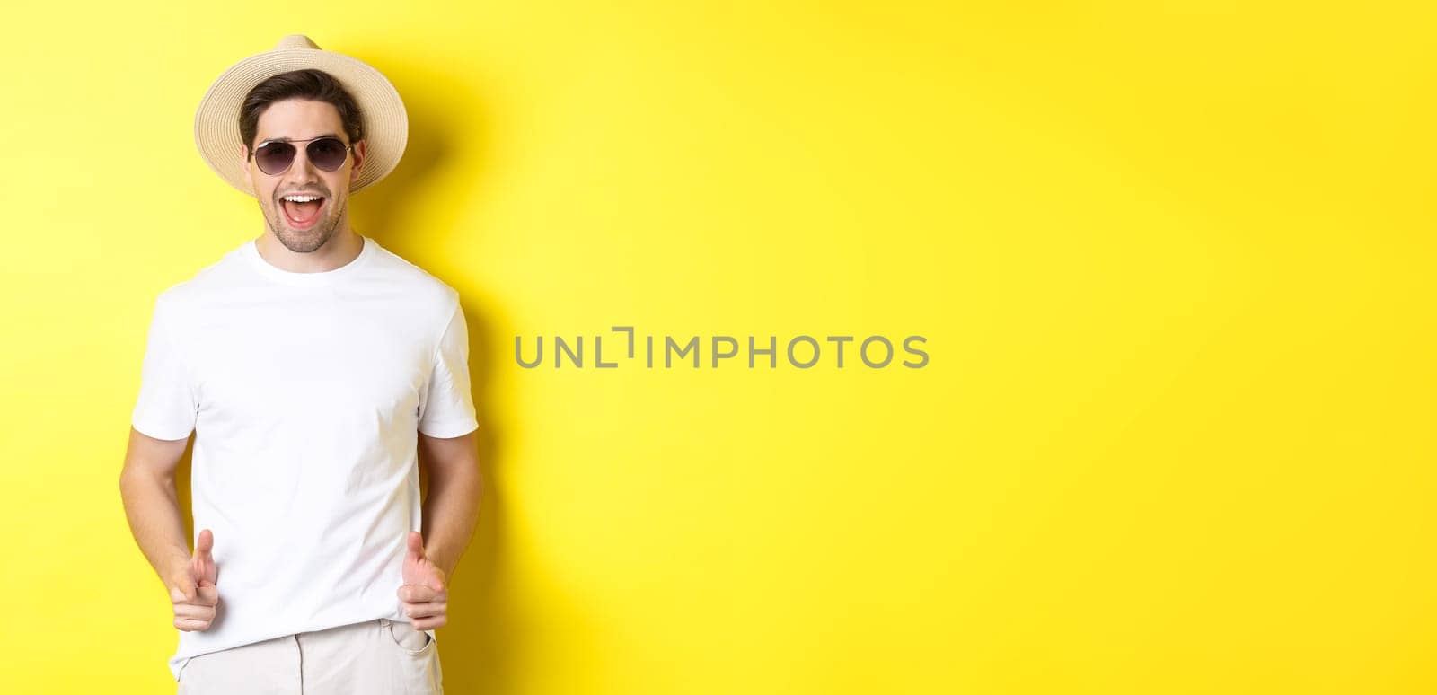 Confident and cheeky guy on vacation flirting with you, pointing finger at camera and winking, wearing summer hat with sunglasses, yellow background.