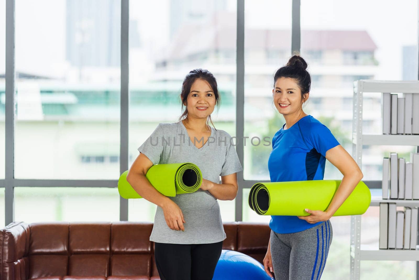 Asian adult and young woman smiling holding a yoga mat after yoga and exercise. Portrait of happy beautiful female standing hold yoga mat indoor studio, sport healthy workout concept