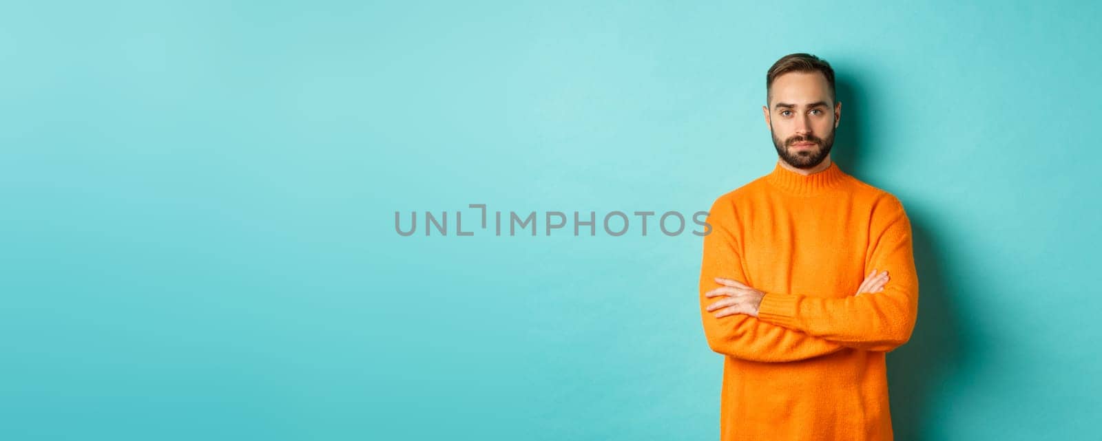 Confident young man looking determined, cross arms on chest, wearing orange winter sweater, standing against turquoise studio background.