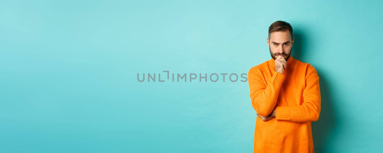 Thougtful young man making decision, looking serious and thinking, choosing, standing near copy space turquoise background.