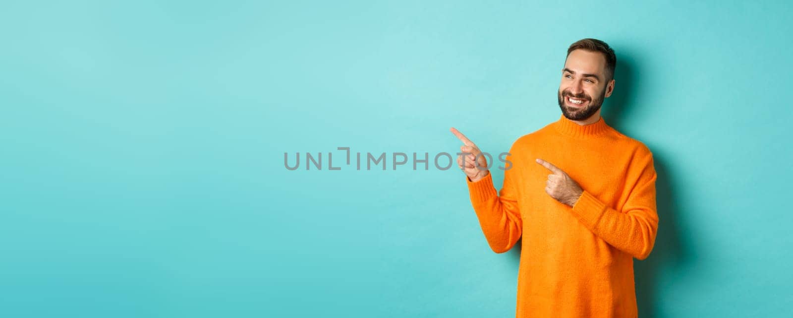 Attractive bearded male model in orange sweater, smiling satisfied and pointing fingers left, standing over turquoise background by Benzoix