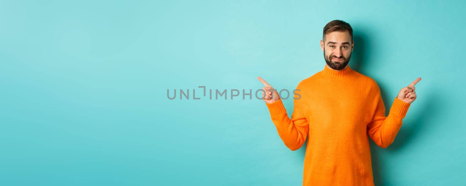 Doubtful and indecisive man pointing fingers sideways, showing different sides, two choices, looking skeptical, standing over light blue background.