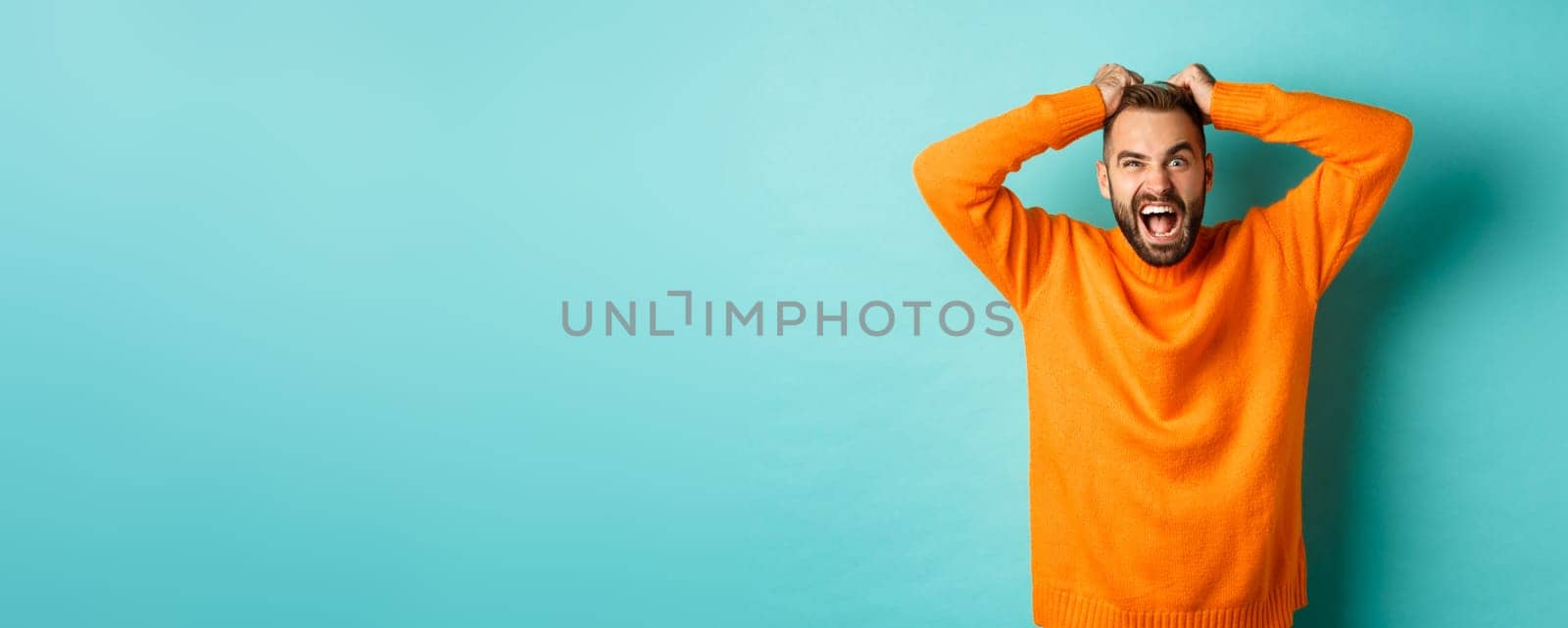 Frustrated man shouting, pulling out hair and screaming angry, losing temper and looking mad, standing over light blue background.