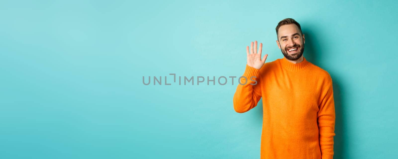 Photo of friendly young man saying hello, smiling and waiving hand, greeting you, standing in orange sweater over light blue background.