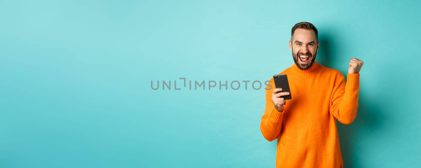 Photo of happy man triumphing, holding mobile phone and make winner gesture, rejoice and achieve goal, standing over light blue background.