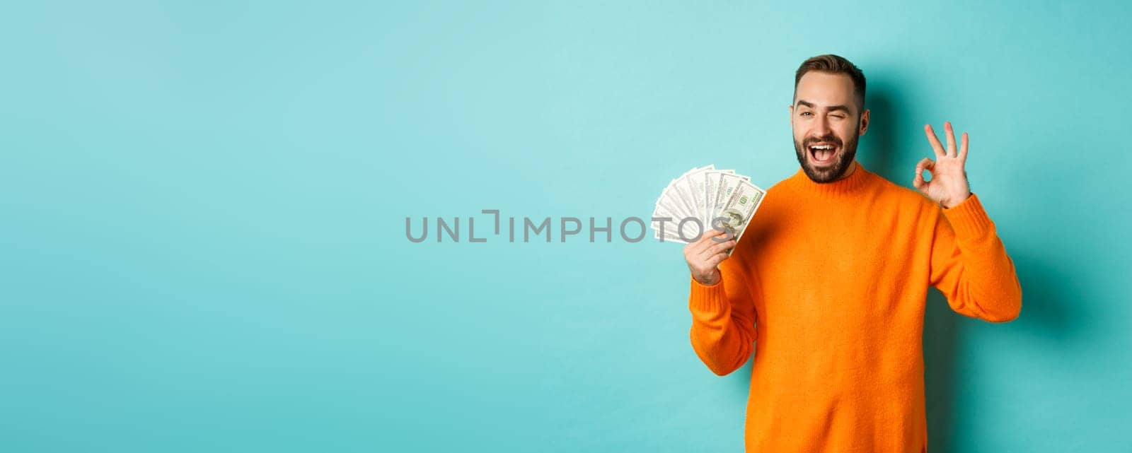 Shopping. Excited guy holding money, showing ok sign and winking, standing over light blue background. Copy space