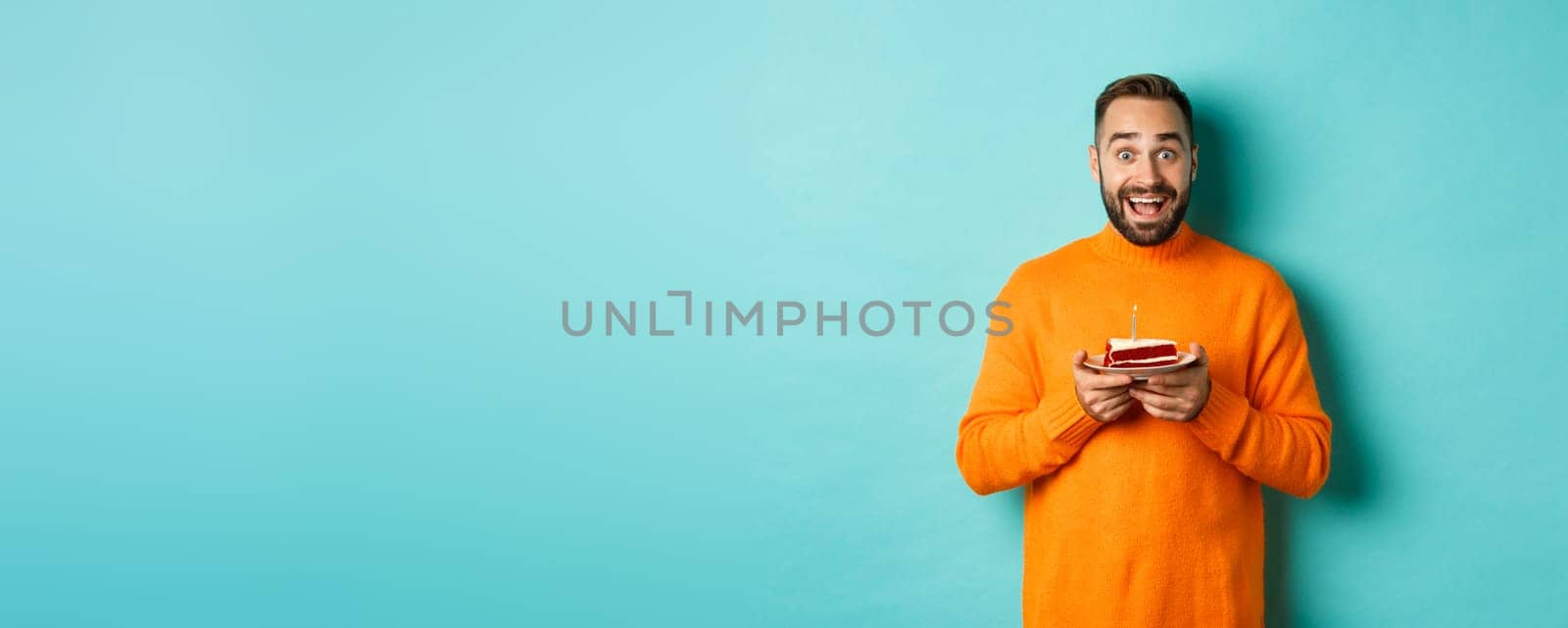 Happy adult man celebrating birthday, holding bday cake with candle and smiling, standing against turquoise background.