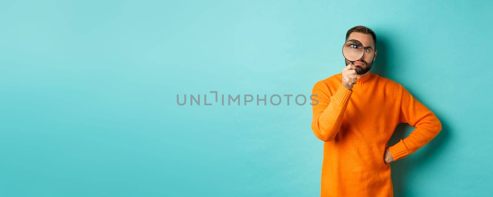 Funny man looking serious through magnifying glass, inspecting something, standing in orange sweater against turquoise background.