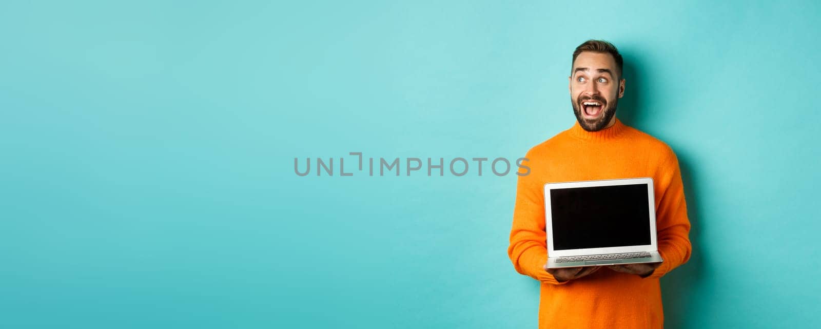 Amazed young man in orange sweater looking at upper left corner, showing laptop screen promo offer, standing over turquoise background.