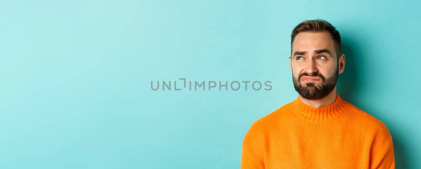 Close-up of handsome caucasian man looking left disappointed and skeptical, staring at logo, wearing orange sweater, standing against turquoise background.