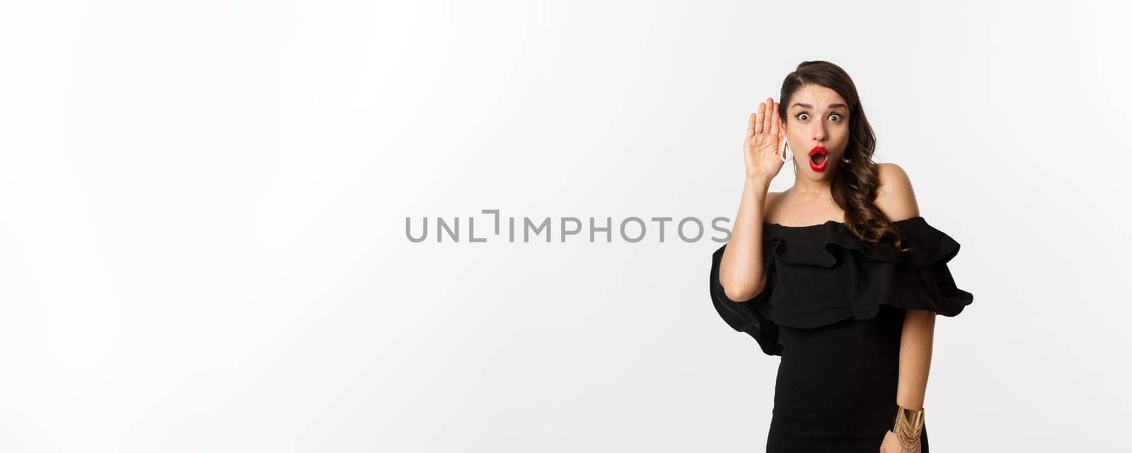 Fashion and beauty. Image of excited woman eavesdropping, holding hand near ear and leaning left to hear gossips, white background.