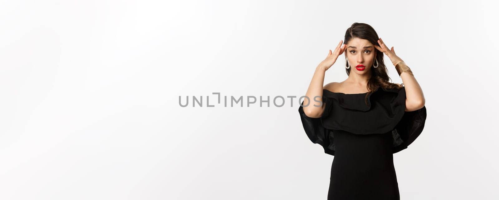 Fashion and beauty. Annoyed and tired woman in black dress, touching head and roll eyes bothered, standing distressed against white background.