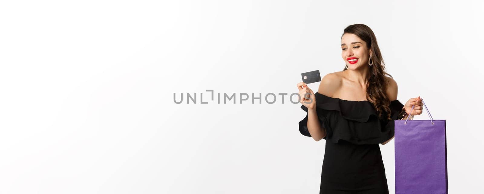 Stylish young woman in black dress going shopping, holding bag and credit card, smiling pleased, standing over white background by Benzoix