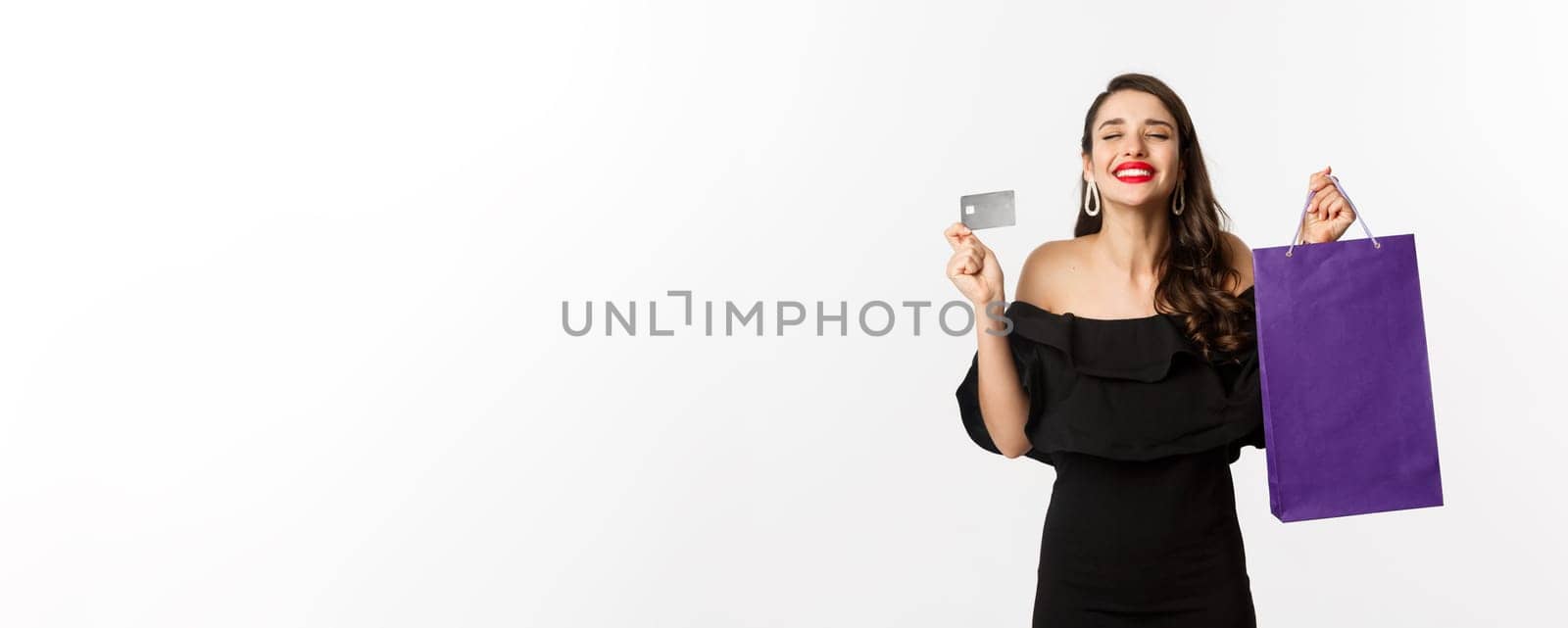 Stylish young woman in black dress going shopping, holding bag and credit card, smiling pleased, standing over white background by Benzoix