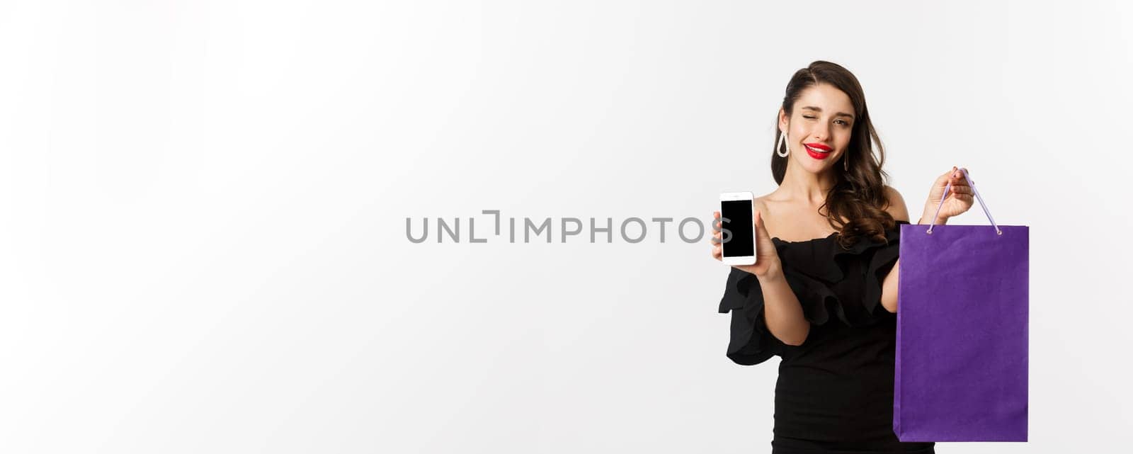 Beauty and shopping concept. Beautiful and stylish woman winking, showing smartphone screen and bag, buying online, standing over white background.