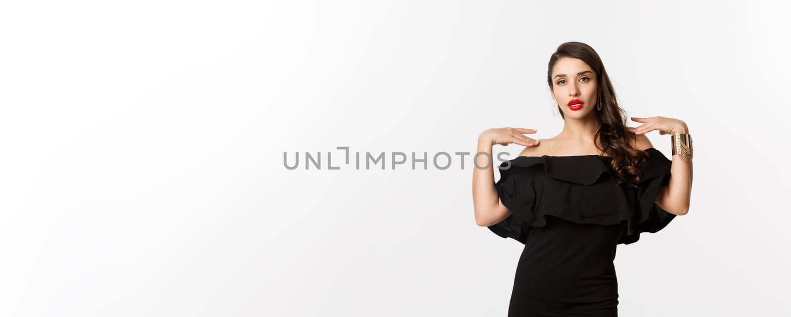 Confident and stylish woman in black elegant dress, looking sassy at camera, standing over white background.