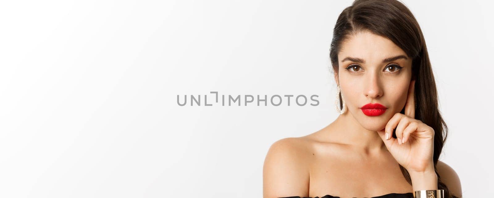 Fashion and beauty concept. Close-up of elegant beautiful woman in black dress, evening makeup and red lipstick, looking sassy at camera, standing over white background.