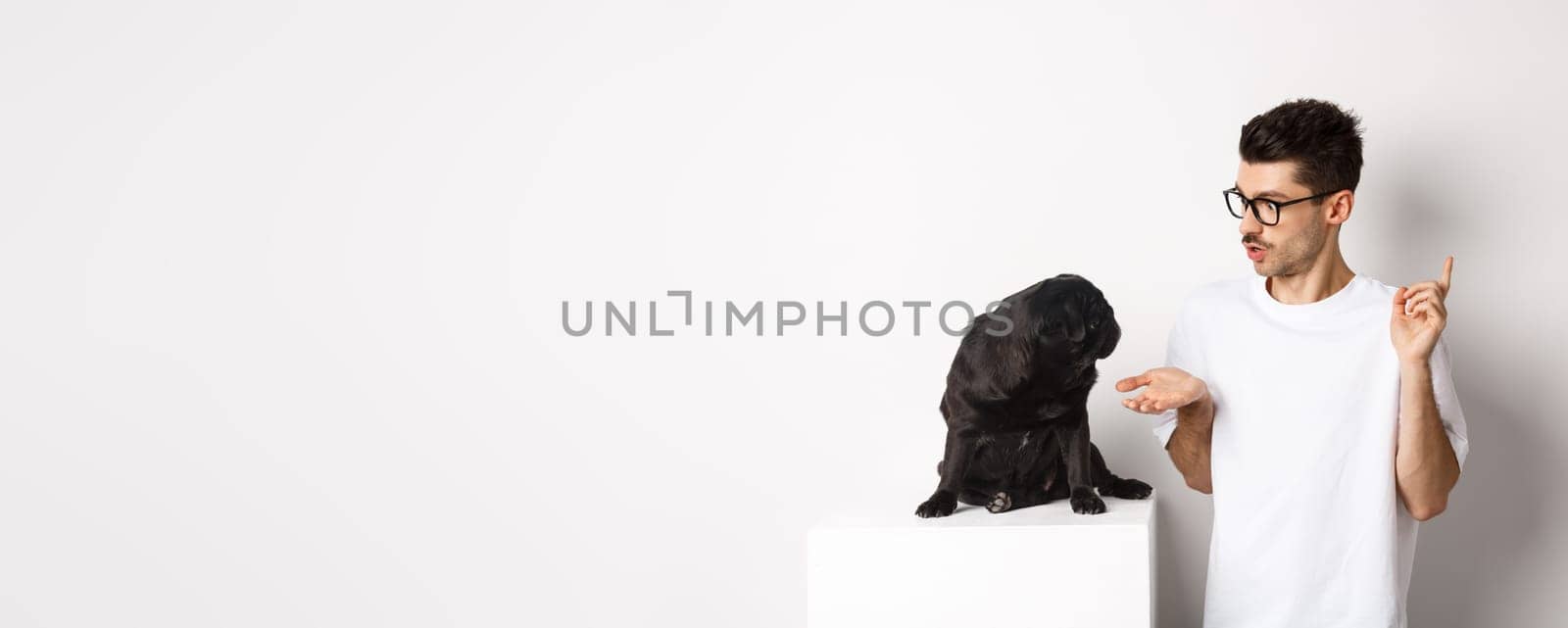 Handsome young man teaching dog commands, talking to cute black pug, standing over white background by Benzoix