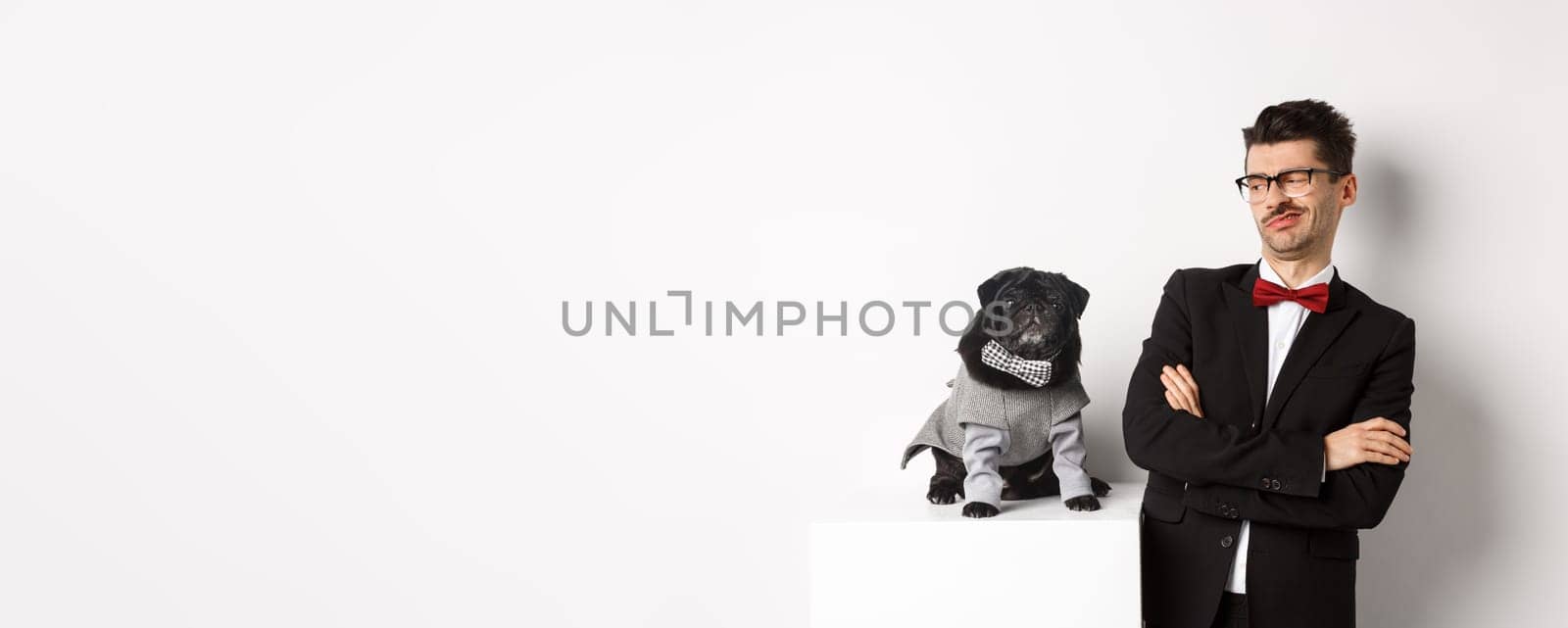 Animals, party and celebration concept. Image of funny young man in suit and glasses, looking skeptical at cute pug in costume, standing over white background by Benzoix