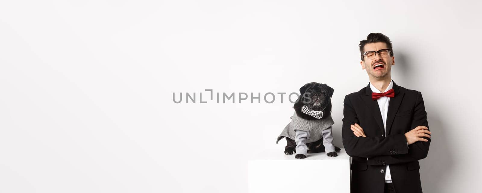 Animals, party and celebration concept. Sad dog owner crying, wearing suit, standing near cute black pug in costume, standing over white background.