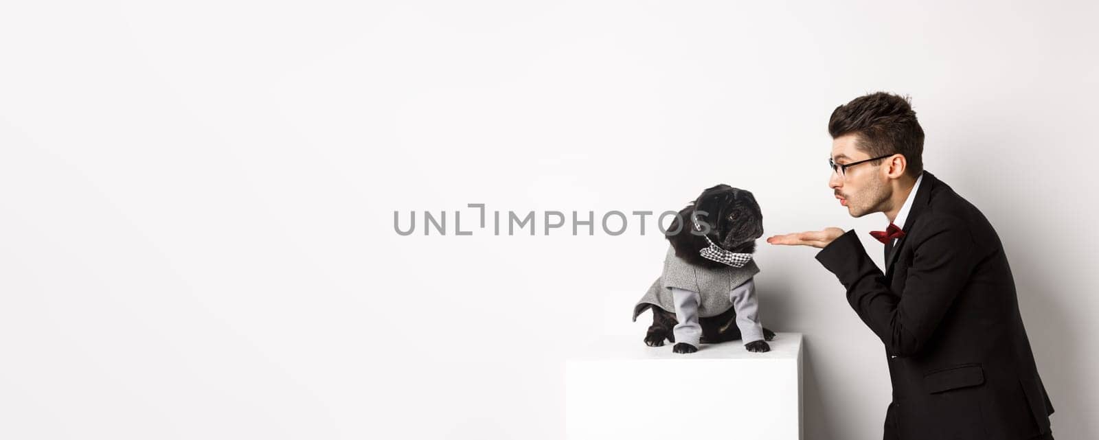 Pets, winter holidays and celebration concept. Handsome young man sending air kiss at cute black puppy wearing costume for New Year, owner standing in suit over white background.