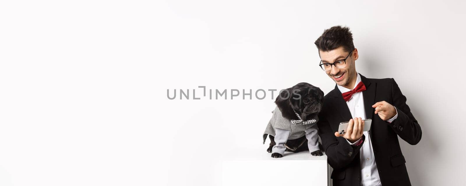 Handsome young man showing something on mobile phone to his dog. Owner shopping online with pet, standing in costumes over white background.