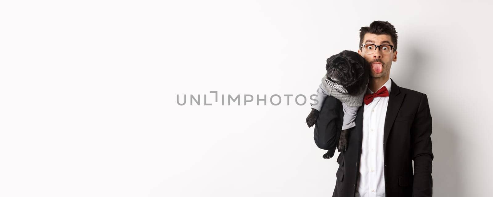 Funny young man in party suit, showing tongue and holding cute black pug on shoulder, celebrating with pet, standing over white background.