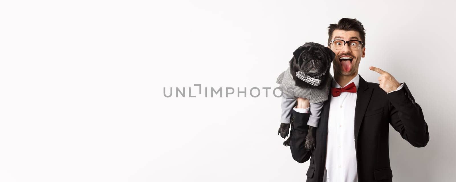 Funny young man in party suit, showing tongue and making grimaces, pointing at cute black dog in winter clothes, standing over white background.
