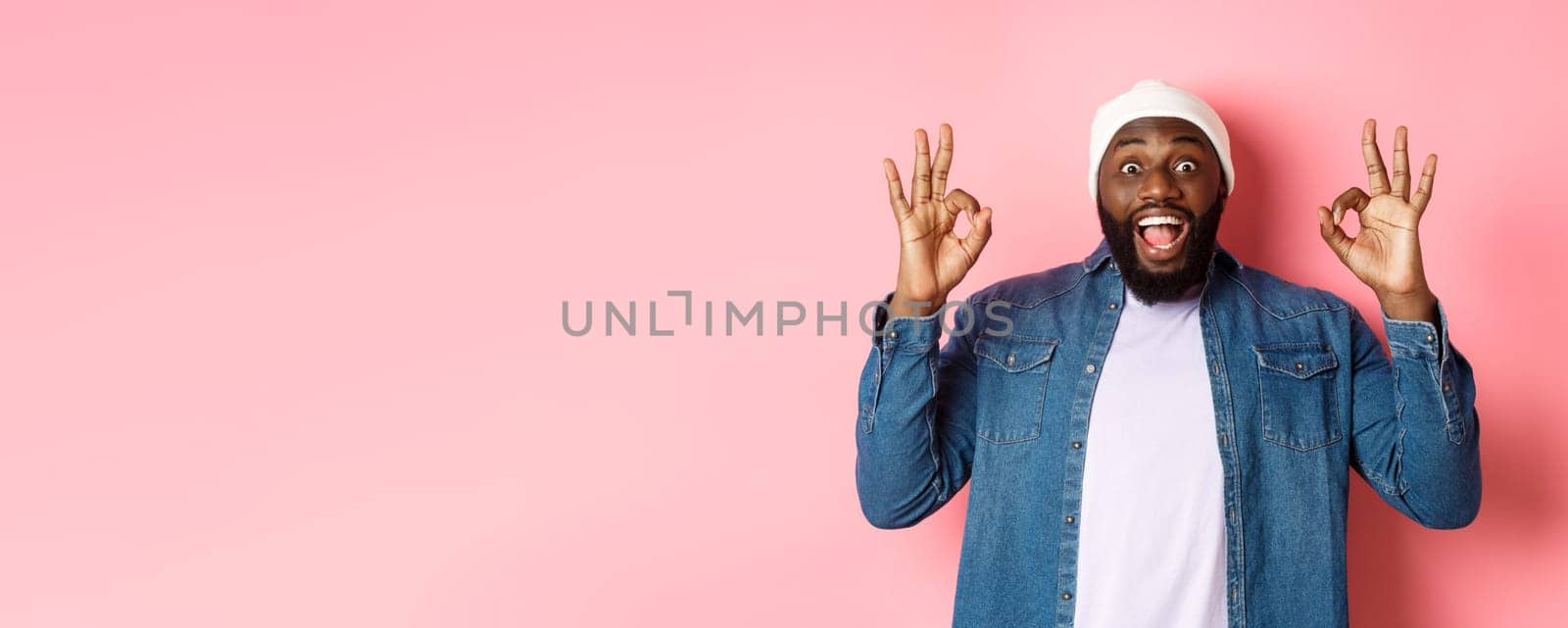 Impressed Black man in beanie and shirt, showing ok signs and staring at camera amazed, smiling happy, praise great offer, recommending product, pink background by Benzoix