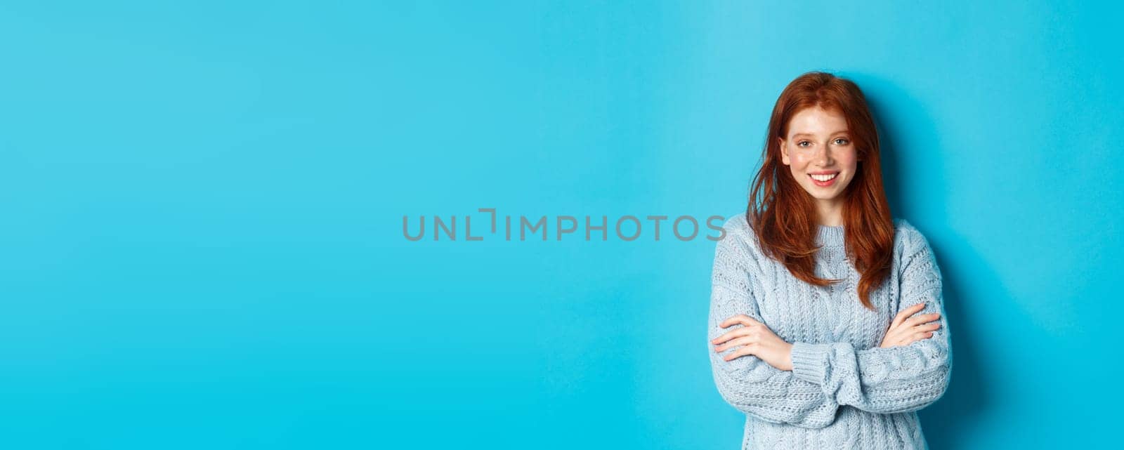 Attractive redhead girl in sweater smiling and staring at camera, standing confident against blue background.