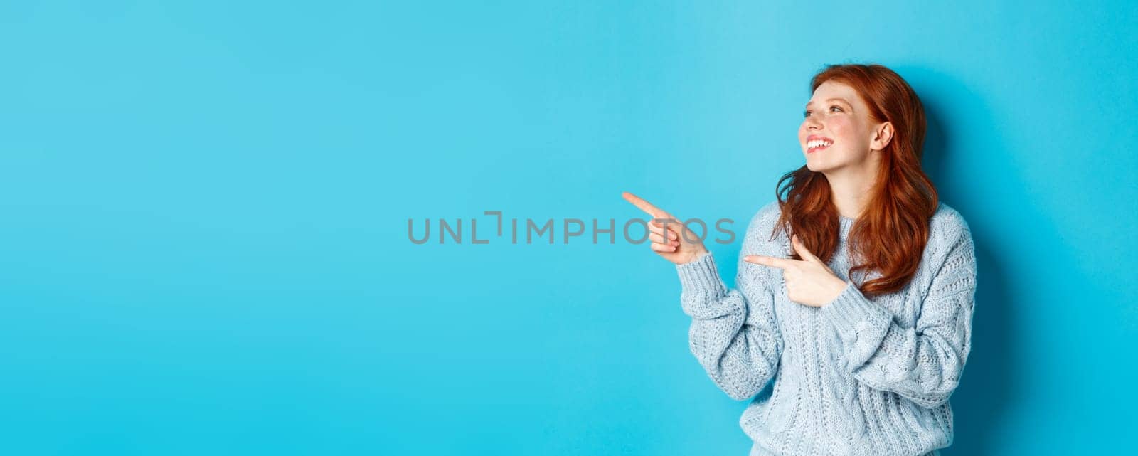 Beautiful happy redhead girl, pointing fingers left and looking at logo pleased, standing in sweater against blue background.