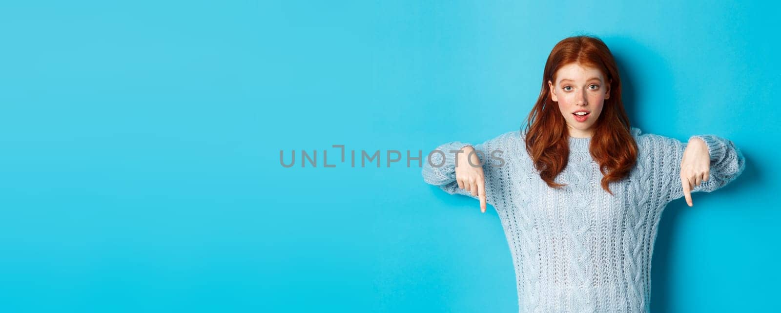 Winter holidays and people concept. Cute teenage girl pointing fingers down, showing advertisement, standing over blue background.