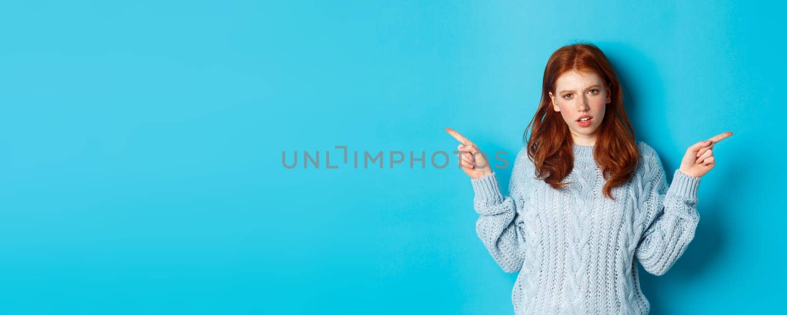 Confused redhead girl in sweater pointing fingers sideways, staring at camera doubtful, standing against blue background. Copy space