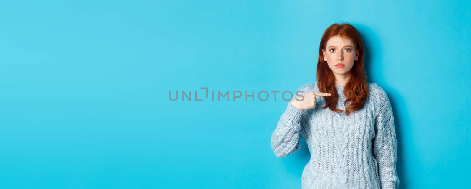 Nervous and confused redhead girl pointing at herself, standing in sweater against blue background by Benzoix