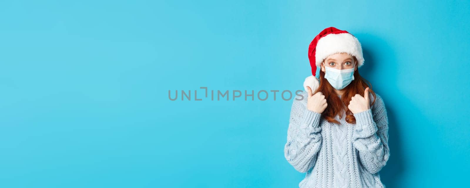 Christmas, quarantine and covid-19 concept. Cute teen redhead girl in santa hat and sweater, wearing face mask from coronavirus, showing thumbs up, standing over blue background.