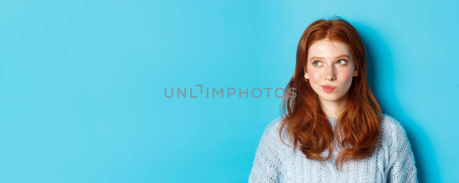 Close-up of cute redhead girl thinking and looking left, have an idea, standing against blue background.