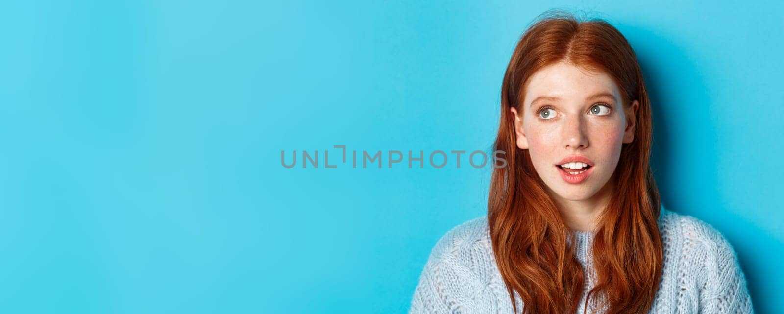 Headshot of thoughtful redhead teen girl looking at upper left corner, staring at logo with curious expression, standing over blue background.