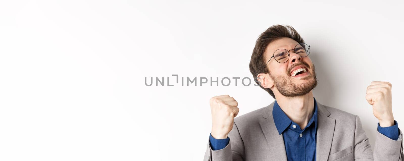 Excited businessman in glasses and suit shouting yes with pleasure and relieved face, shaking fists up, triumphing, winning bet, standing on white background.