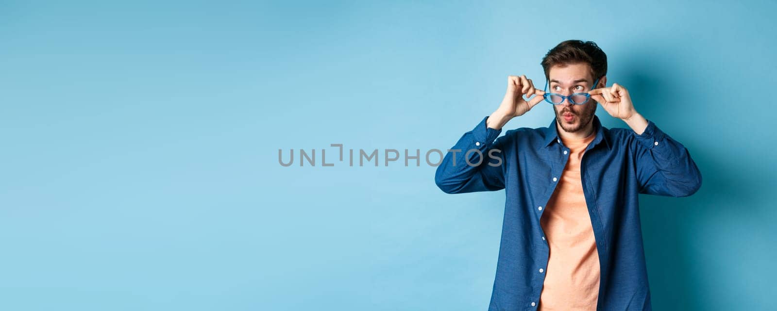 Image of caucasian guy takes-off sunglasses and looking aside with surprised face, checking out empty space for logo, standing on blue background.