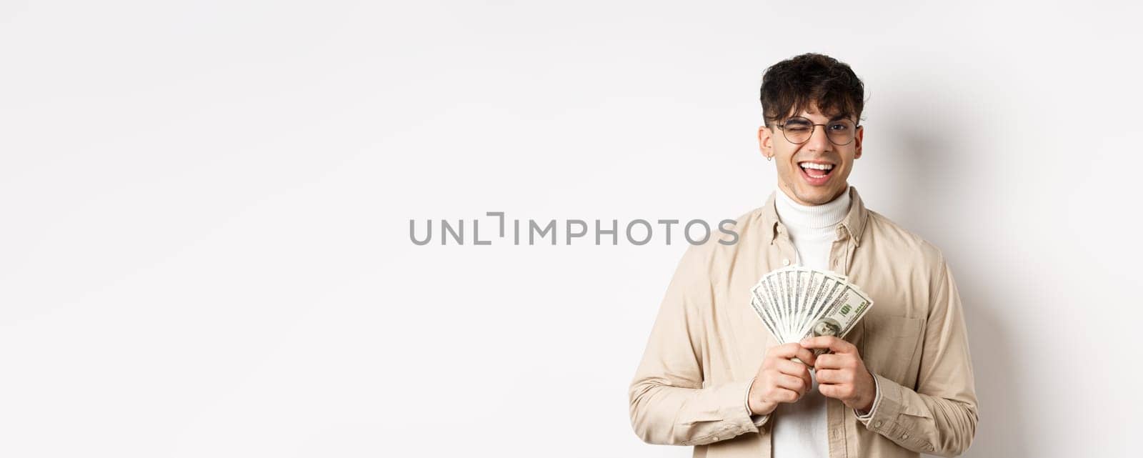 Handsome young man winking and showing dollar bills, holding fan of money banknotes and smiling pleased, making money, standing on white background.