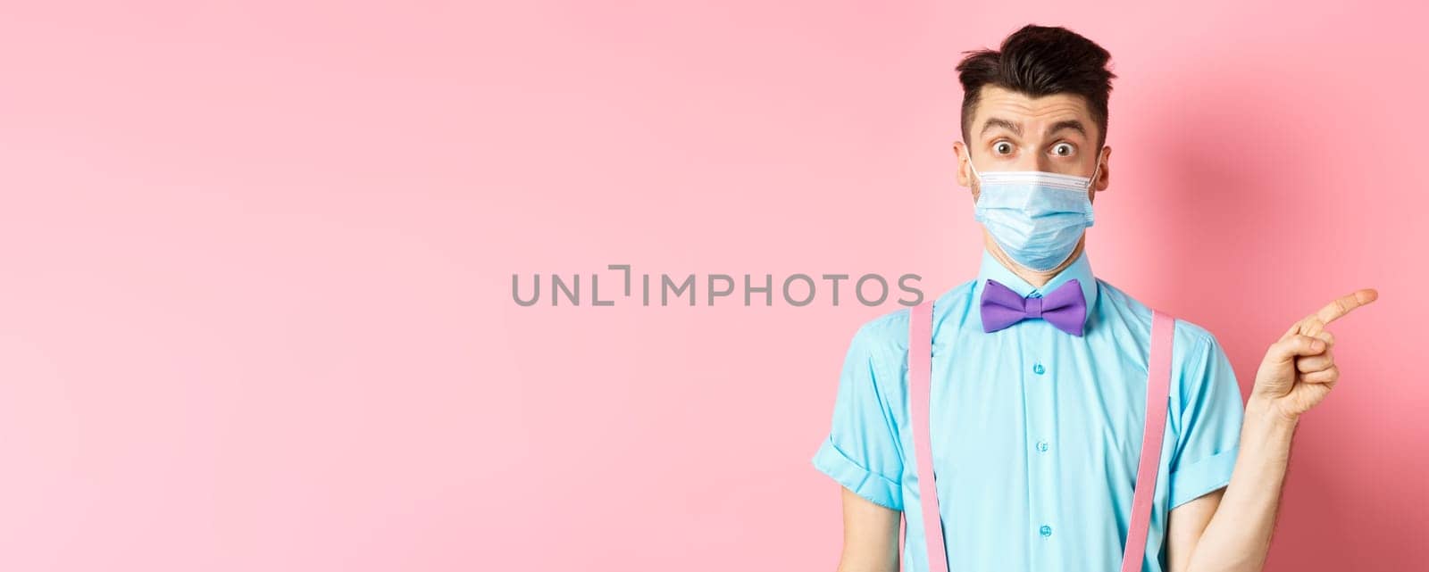 Coronavirus, healthcare and quarantine concept. Man looking surprised in medical mask, asking question and pointing right, curious about promo offer, standing on pink background.