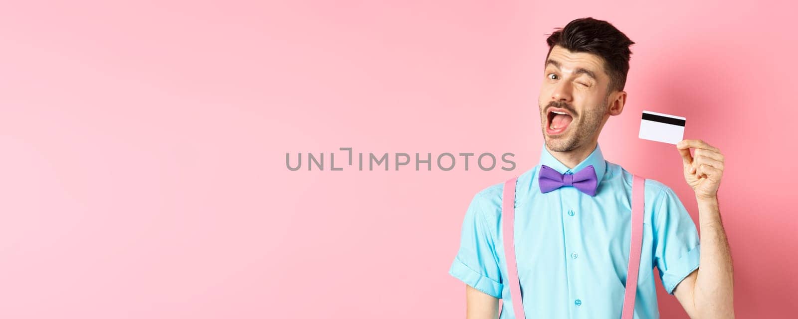 Shopping concept. Funny guy with moustache winking at camera, showing plastic credit card, recommending bank promo offer, standing on pink background.