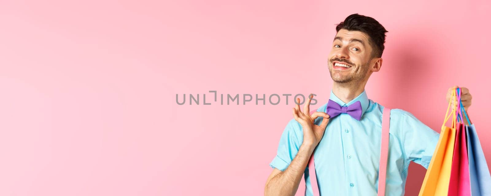 Confident smiling guy adjusting his bow-tie and showing gifts in shopping bags, looking happy, standing over pink background.