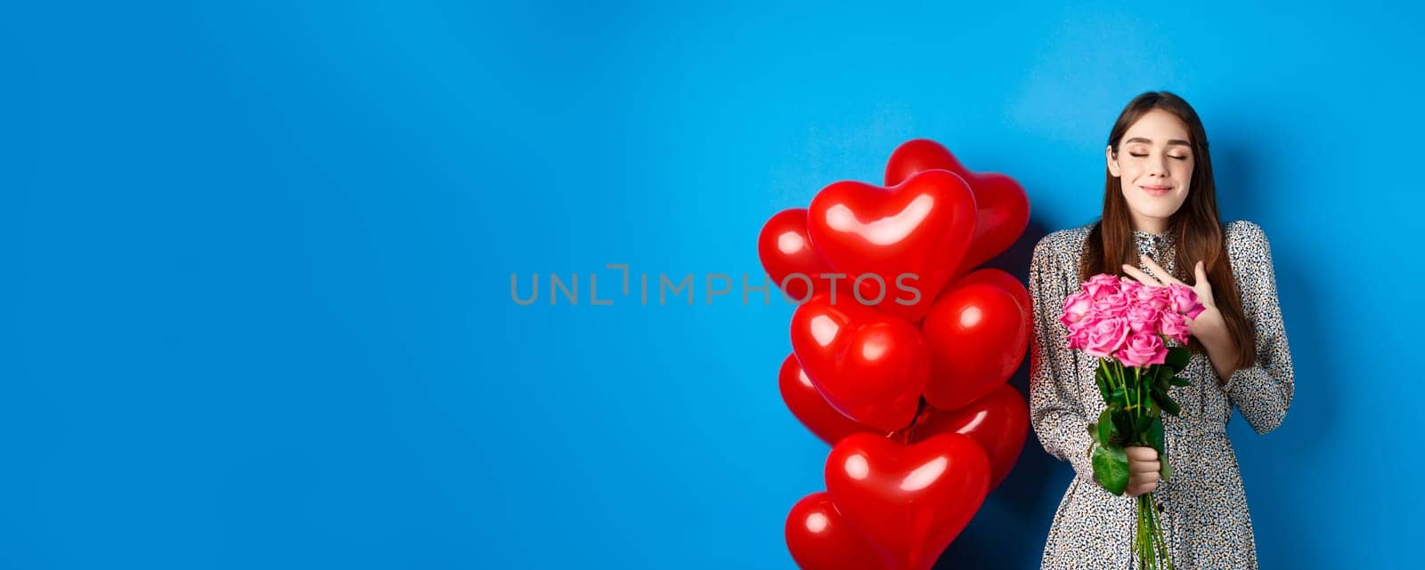 Valentines day. Romantic lovely woman in dress, close eyes and smiling, receiving flowers from lover, smelling bouquet of pink roses, blue background by Benzoix