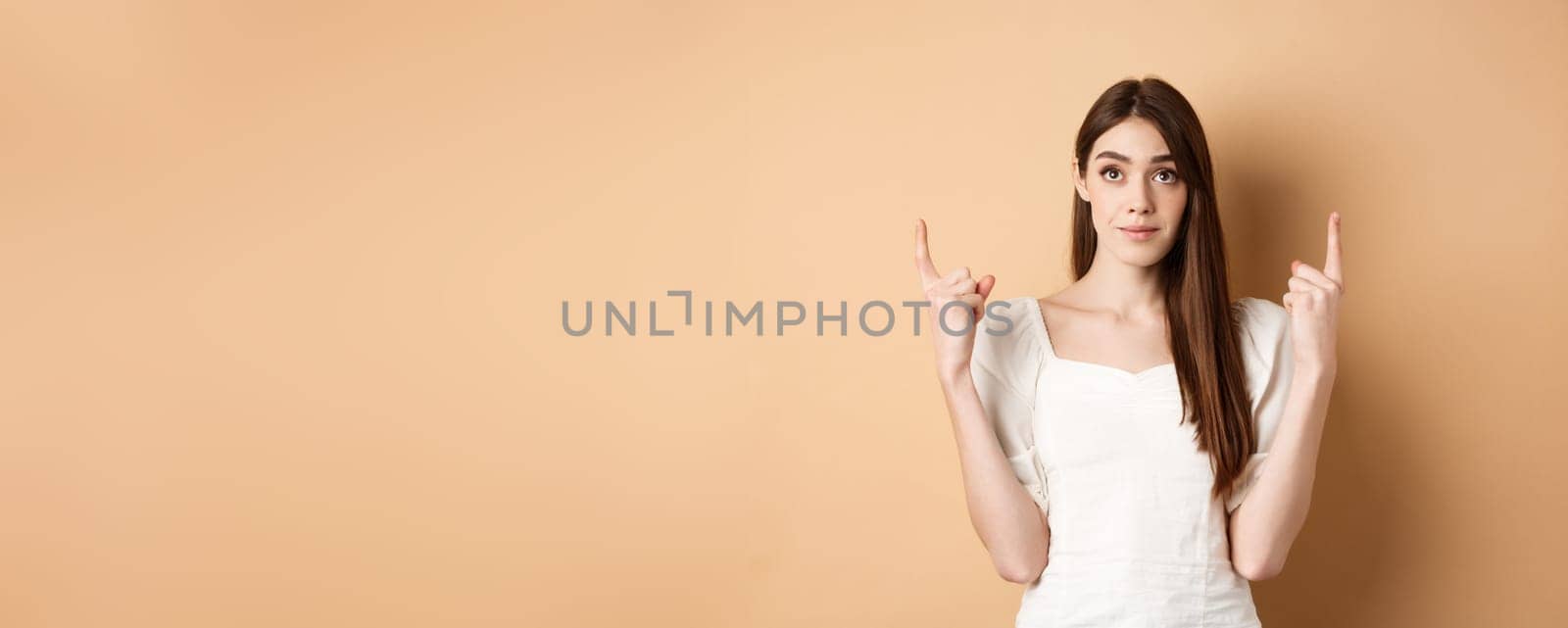 Hopeful romantic girl in white dress pointing fingers up, showing empty space for company logo, standing on beige background by Benzoix