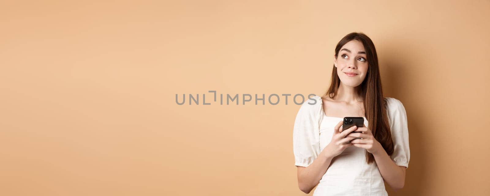 Romantic girl thinking with phone, looking aside and smiling dreamy, using dating app on smartphone, standing on beige background.