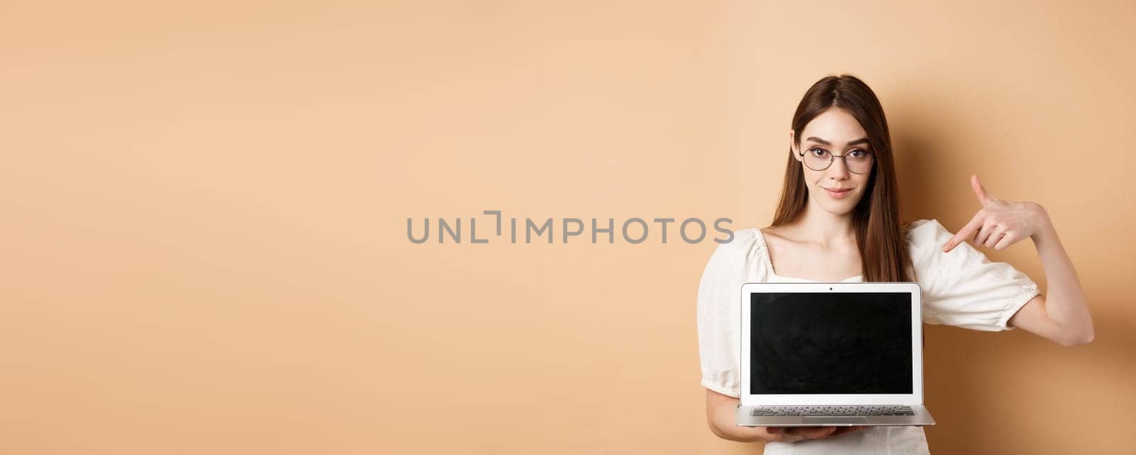 Confident young woman in glasses pointing at laptop screen, showing computer display, standing on beige background.