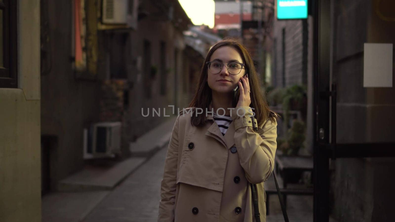 A young woman is talking on the phone and walking along a narrow street. A girl with glasses walks on the street with a phone. 4k