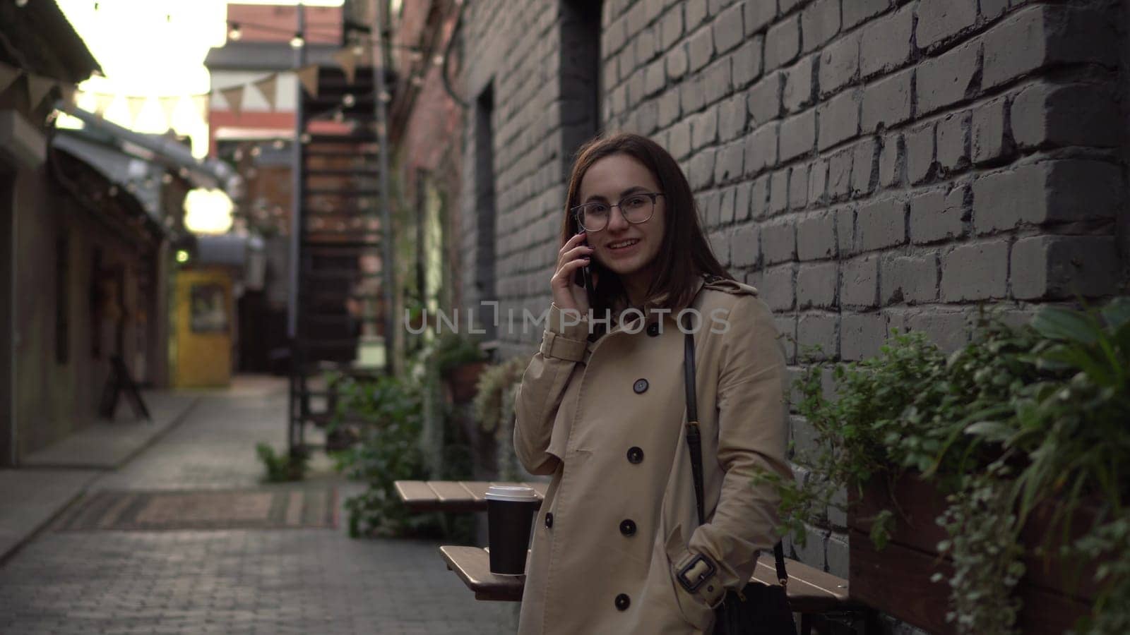 A young woman talks on the phone while standing against a wall on a narrow street. A girl with glasses and a coat speaks on the phone. 4k